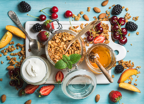 Healthy breakfast ingredients. Oat granola in open glass jar, yogurt, fruit, berries, honey and mint on white ceramic board over blue background, top view