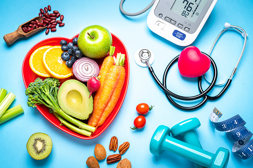Healthy lifestyle concepts: red heart shape plate with fresh organic fruits and vegetables shot on blue background. A digital blood pressure monitor, doctor stethoscope, dumbbells and tape measure are beside the plate  This type of foods are rich in antioxidants and flavonoids that prevents heart diseases, lower cholesterol and help to keep a well balanced diet. High resolution 42Mp studio digital capture taken with SONY A7rII and Zeiss Batis 40mm F2.0 CF lens