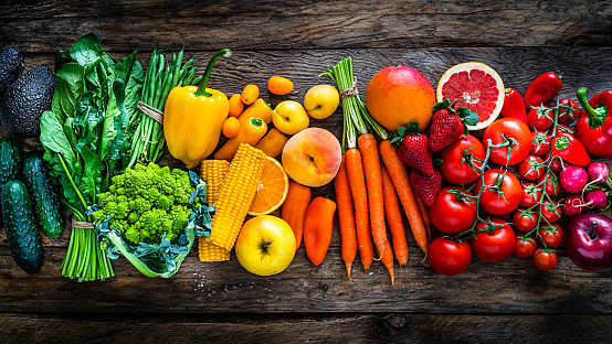 Top view of a large assortment of healthy fresh rainbow colored organic fruits and vegetables arranged side by side on rustic wooden table. The composition includes carrots, onion, tomatoes, avocado, corn, green bean, cucumber, broccoli, spinach, apples, strawberries, mango, grape fruit, peach, oranges, bell pepper, radish among others. The composition is at the left of an horizontal frame leaving useful copy space for text and/or logo at the right. High resolution 42Mp studio digital capture taken with SONY A7rII and Zeiss Batis 40mm F2.0 CF lens