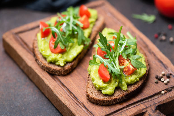 Healthy toast with avocado, tomato, arugula Healthy vegan toast with avocado, tomato, arugula on wooden serving board, closeup view, horizontal image. Snack, lunch or vegan breakfast toasted bread stock pictures, royalty-free photos & images