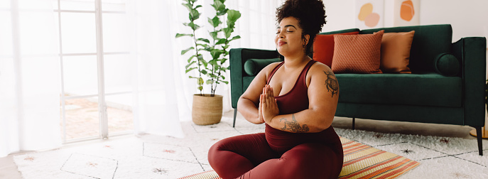 Healthy female in practicing meditation yoga at home. Body positive woman exercising with eyes closed and hands joined while sitting cross legged in living room.