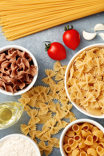 Variety of raw pasta in different types and shapes with tomatoes, oil, garlic and flour. Overhead view.