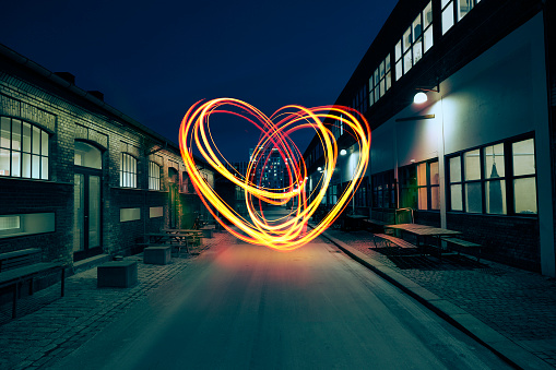 With light, a heart is painted in the air in the middle of a small street between two industrial buildings.