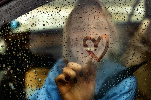 Happy woman drawing heart on car window during rainy day