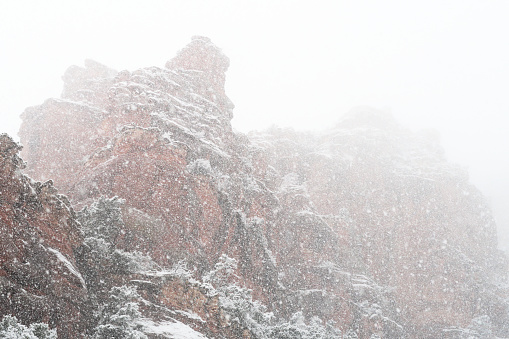 Large blizzard snow flurries obscure red rock cliffs in Sedona, Arizona.  The scene is entirely filled with large snowflakes.  The red rock cliffs with areas of dark green high desert vegetation occupy the bottom two thirds of the horizontal frame, snow-filled sky the upper third.  The cliffs appear slightly more clearly at the foreground, left of frame, dropping further into obscurity in the background at right of frame.