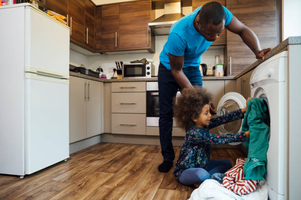 A Helping Hand Wide shot of a mixed race girl sitting on the floor in the kitchen and helping her father put the washing into the washing machine. laundry photos stock pictures, royalty-free photos & images