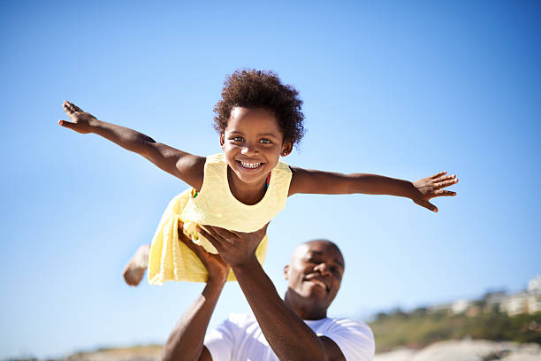 Helping his daughter soar! A father lifting his adorable daughter into the air while enjoying a day at the beach african american children stock pictures, royalty-free photos & images