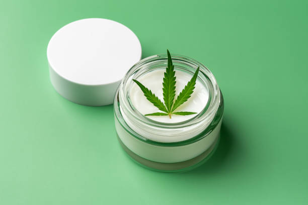 Hemp skin care moisturizer and fresh cannabis sativa leaf in an open glass jar  on a green background. Hemp herbal cosmetics and medical marijuana plant for face and body care concepts. stock photo