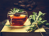 Herbal tea made from sage in glass cup standing on books, nearby lies a bundle of sage over dark wooden background. Retro toned.