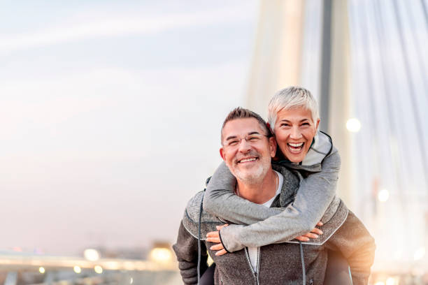 Photo of Happy mature couple having fun, hugging in the city on a autumn day. Love story true feelings concept. Portrait of friendly peaceful fitness couple in love. Couple enjoying the outdoors together. Sporty couple in love enjoying each other. Handsome  man giving a gray hair woman a piggy back ride.