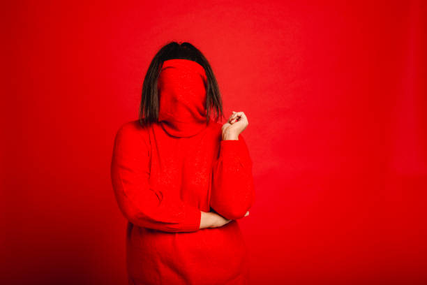 Hiding in Plain Sight Portrait of a young female adult using her turtleneck to cover her face. She is posing with her handoff her chin while standing infront of a red studio background. shy photos stock pictures, royalty-free photos & images