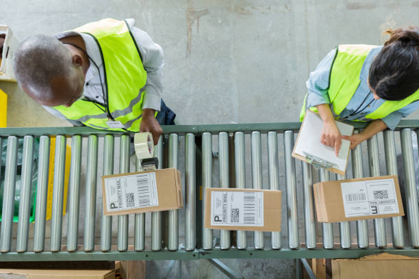 High angle view of people working in distribution warehouse Male and female distribution warehouse employees process customers' orders. The male employee is taping a box. The female employ is writing on a clipboard. conveyor belt stock pictures, royalty-free photos & images