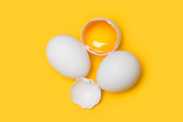 High angle view of small group of white raw eggs, one is broken on yellow background stock photo