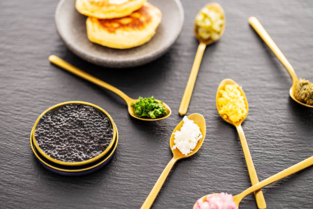 High quality real black sturgeon caviar in a golden tin can aside blinis in a small plate and golden spoons with pickles and herbs on a slate background. Side view. stock photo