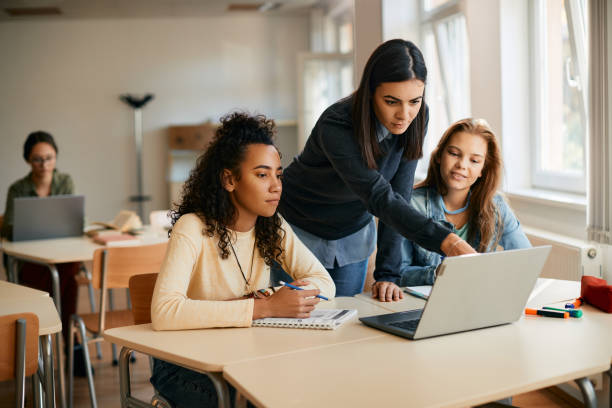 High school professor assisting her students in e-learning on laptop in the classroom. stock photo