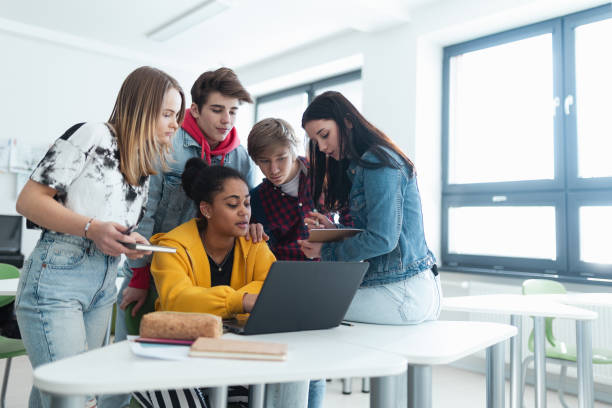 High school students sitting together at desk and using laptop and talking during break in classroom. stock photo