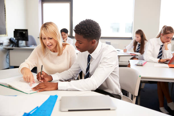 High School Tutor Giving Uniformed Male Student One To One Tuition At Desk In Classroom High School Tutor Giving Uniformed Male Student One To One Tuition At Desk In Classroom high school teacher stock pictures, royalty-free photos & images