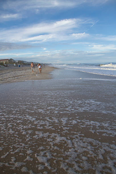 High Tide and Beachcombers Carolina Beach, Pleasure Island, North Carolina, USA - April 18, 2015: The foamy tide comes in covering the beach in front of people walking along the ocean with blue sky and bright puffy clouds overhead carolina beach north carolina stock pictures, royalty-free photos & images
