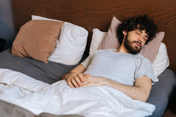 High-angle view of handsome bearded young man sleeping peacefully lying on back in large comfortable double bed under white blanket. stock photo