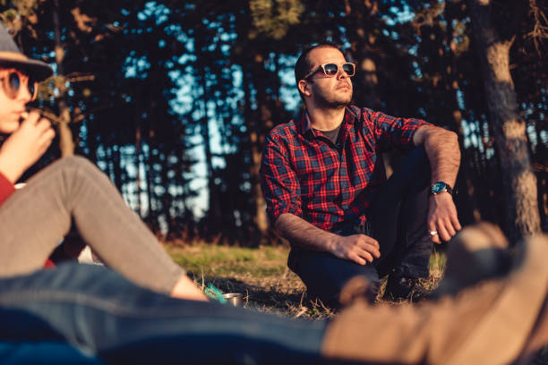 Hiker resting on a grass with friends and enjoying sunset Hiker wearing plaid shirt and sunglasses resting on a grass by the pine forest with friends and enjoying looking at sunset plaid shirt stock pictures, royalty-free photos & images
