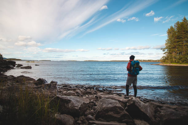 Hiker wearing a jacket and carrying a backpack walks along the beach and watches Lake Jatkonjarvi at sunset in Koli National Park, eastern Finland. Man aged 24 wearing sports clothes. Active lifestyle stock photo