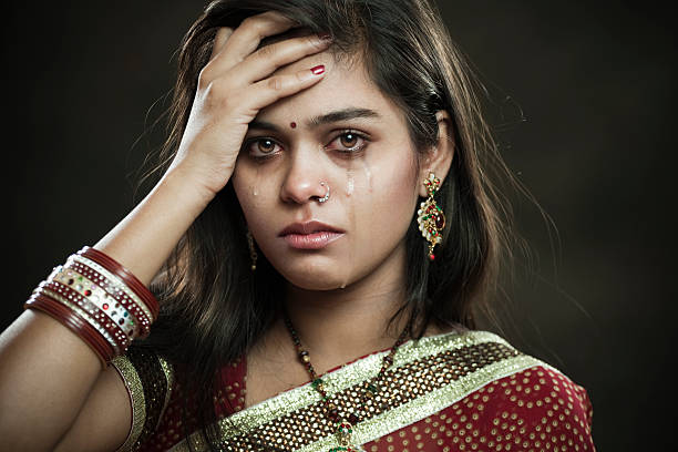 Hindu married woman crying with tears and looking at camera. Indoor isolated image shot against dark background of a newly married sad Hindu young woman of Indian ethnicity crying with tears while holding her head in hand. She is in traditional Hindu dress which is sari, blouse, bangles, necklace and earrings. Horizontal composition with copy space and selective focus. indian bride stock pictures, royalty-free photos & images