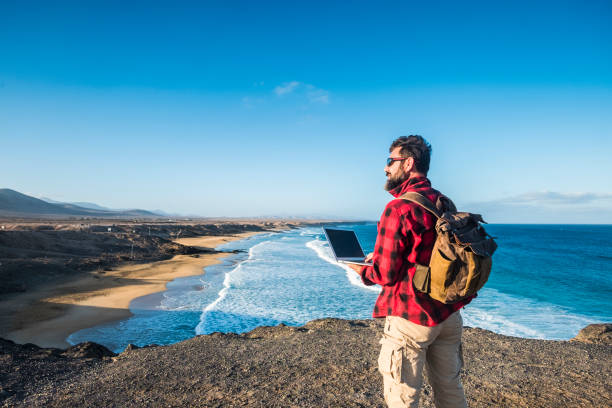 hipster people using outdoor cimputer laptop with internet connection for work or to communicate at home - traveler with backpack and alternative travel vacation - beach and sea in background - cargo canarias imagens e fotografias de stock