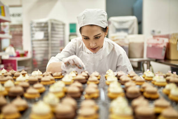 Hispanic American Female Baker Decorating Vegan Cupcakes Low angle view of focused baker in mid 30s decorating fresh batch of vegan cupcakes in commercial kitchen. artisanal food and drink photos stock pictures, royalty-free photos & images