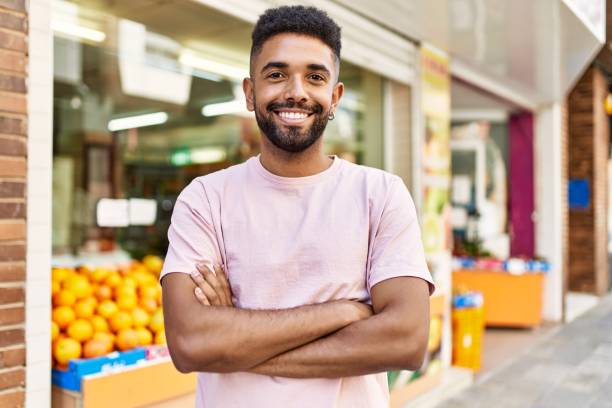 Hispanic man standing by fruits and vegetables shop. Smiling happy with crossed arms by marketplace stock photo