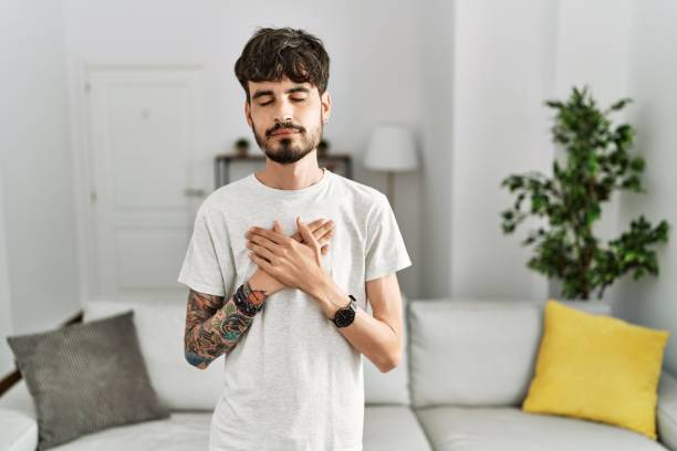 Hispanic man with beard at the living room at home smiling with hands on chest with closed eyes and grateful gesture on face. health concept. stock photo