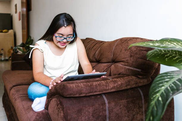Hispanic teen girl with down syndrome using a tablet while sitting on the sofa at home, in disability concept in Latin America stock photo