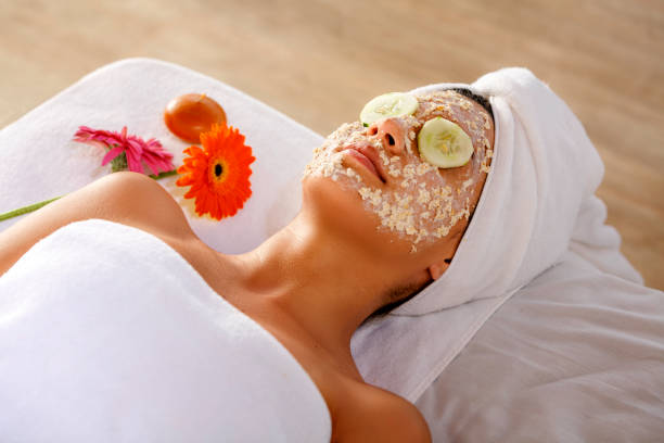 Hispanic young woman laying on bed and getting oatmeal face scrub and resting with cucumber slices on eyes. stock photo