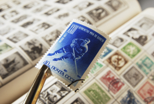 Close-up of unrecognizable senior adult man inserting postage stamps at collection book sitting at wooden table, selective focus. Mature aged man looking album with old stamps philately at home.
