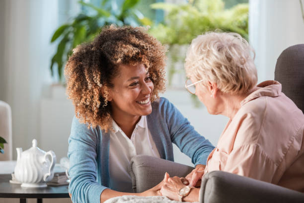 Home caregiver taking care of elderly woman Smiling young female caregiver holding hands and talking with senior woman in living room healthcare worker stock pictures, royalty-free photos & images