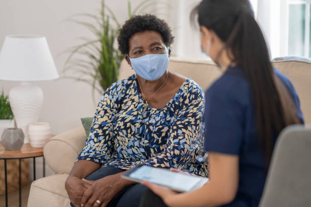 Home caregiver visiting with patient wearing masks Female nurse visiting senior patient in her home during the COVID-19 pandemic, both are wearing masks to avoid the transfer of germs. home caregiver stock pictures, royalty-free photos & images