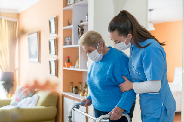 Home caregiver wearing a facemask while assisting a senior woman walking with a mobility walker Home caregiver wearing a facemask while assisting a senior woman walking with a mobility walker during the COVID-19 pandemic - assisted living concepts elderly care stock pictures, royalty-free photos & images