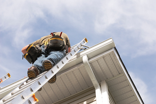 A handyman repairs his rain gutters.  He is up a ladder, photo taken from ground looking up, low angle view.  He wears a tool belt, sky and clouds, good copy space.