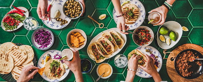Family or friends home taco party. Flat-lay of Mexican traditional dishes Tacos with beef meat, corn tortillas , tomato salsa and peoples hands over green background, top view. Mexican cuisine concept