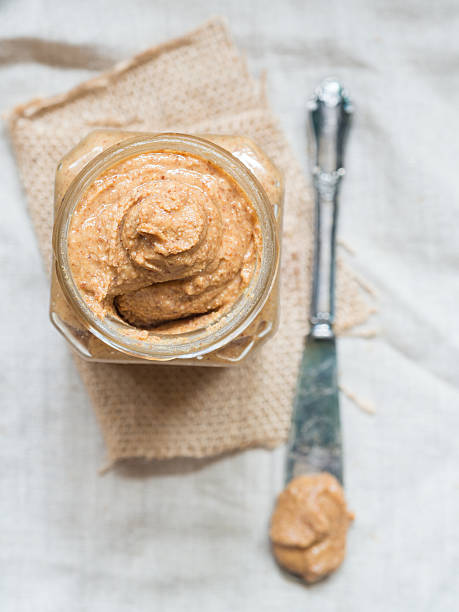 Homemade natural almond butter in a jar, view from above Vertical photo of homemade natural almond butter in a glass jar placed on rustic beige table cloth and with a butter knife on the side. Pastel colors. View from above. almond butter stock pictures, royalty-free photos & images