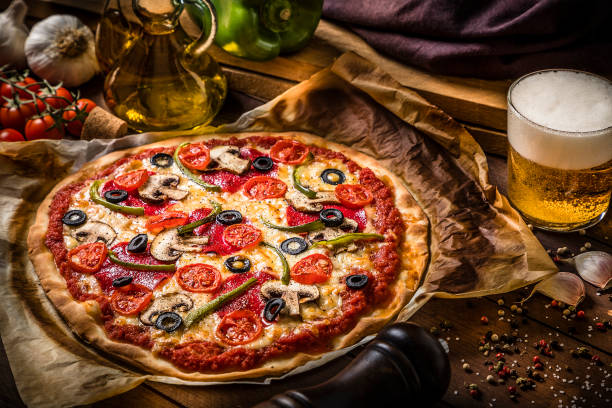 Homemade pizza with beer High angle view of a homemade pizza  made with salami, mozzarella, mushrooms, cherry tomatoes, black olives and green bell pepper alongside a drinking glass full of beer and some ingredients. Low key DSLR photo taken with Canon EOS 6D Mark II and Canon EF 24-105 mm f/4L dough photos stock pictures, royalty-free photos & images