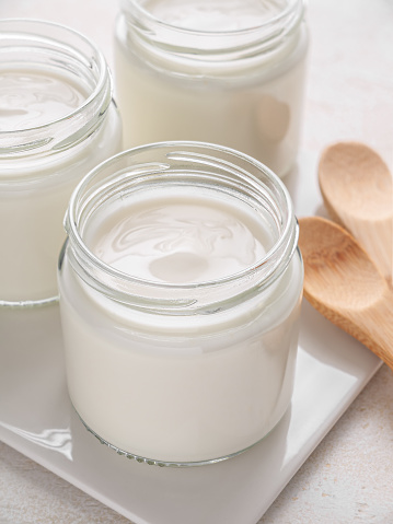 Glass jars of homemade plain yogurt with wooden spoons on white tray. Vertical. Close up.