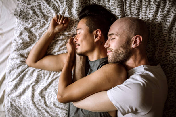 Homosexual partners sleeping in bed together Directly above of gay partners in bed, lying face to face and, looking each other and cuddling gay spooning stock pictures, royalty-free photos & images
