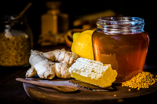 Front view of a honey jar surrounded by lemon, ginger, a honeycomb, a honey dipper and bee pollen. Objects are on a rustic cutting board and against black background. Low key DSLR photo taken with Canon EOS 6D Mark II and Canon EF 24-105 mm f/4L