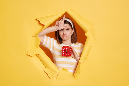 Horizontal shot of sad Caucasian woman wearing hair band and striped t shirt standing in yellow paper hole, holding in hand red alarm clock and showing looser gesture.