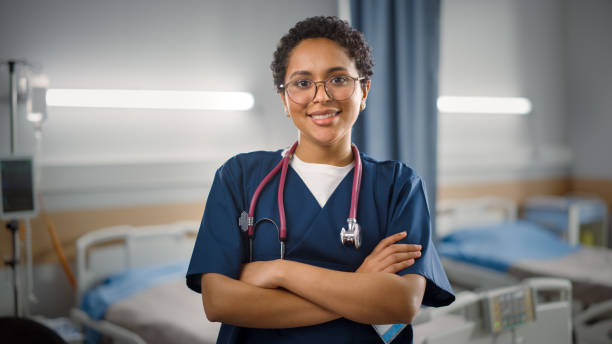 Hospital Ward: Portrait of Posing Beautiful Black Female Head Nurse, Doctor, Surgeon Smiles Charmingly and Kindly Looks at Camera. Modern Clinic with Advanced Equipment and Professional Staff stock photo