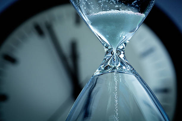 Hourglass Sands of Time Deadline A hourglass with falling sand in front of a clock reaching midnight. Concept photo urgency, and time is running out and deadline is approaching. Close-up of hour glass is photographed in horizontal format with copy space, against a soft-focus clock face in the background. deadline stock pictures, royalty-free photos & images