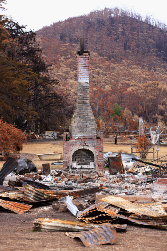 The remains of a house destroyed by a catastrophic bushfire.  The chimney stands stark against the backdrop of a charred hillside. In the foreground is a jumble of burnt and twisted roofing iron.