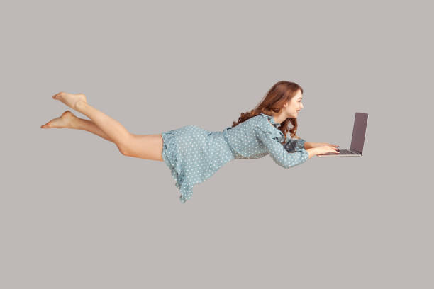 Hovering in air. Happy cheerful girl ruffle dress levitating with laptop, typing keyboard, reading good news message Hovering in air. Happy cheerful girl ruffle dress levitating with laptop, typing keyboard, reading good news message on computer while flying in mid-air. studio shot isolated on gray background hovering stock pictures, royalty-free photos & images
