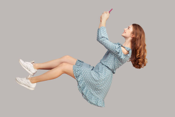 Hovering in air. Smiling girl ruffle dress levitating with mobile phone, reading message chatting happy joyful Hovering in air. Smiling girl ruffle dress levitating with mobile phone, reading message chatting happy joyful in social network online, surfing web while flying. indoor studio shot isolated on gray levitation stock pictures, royalty-free photos & images