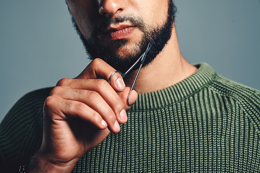 Cropped studio shot of a young man trimming his beard against a grey background